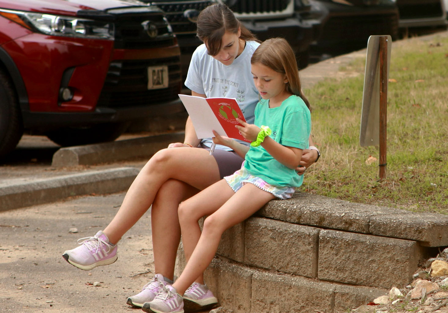 A young mother and her elementary age daughter reading a Christian devotion booklet together outside.