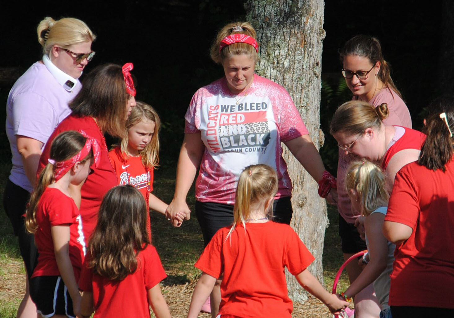 A group of mothers and daughters wearing red t-shirts and holding hands in a circle outside.